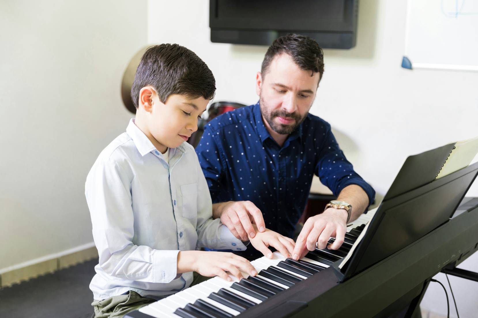 Un bambino con gli occhi chiusi davanti a una pianola viene aiutato da un uomo con barba e camicia blu